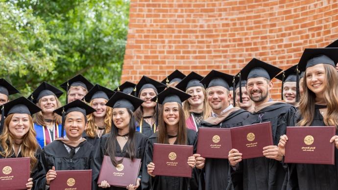 毕业生们头戴帽子，身穿学士服，在默奇森塔前摆姿势拍照, holding their degrees
